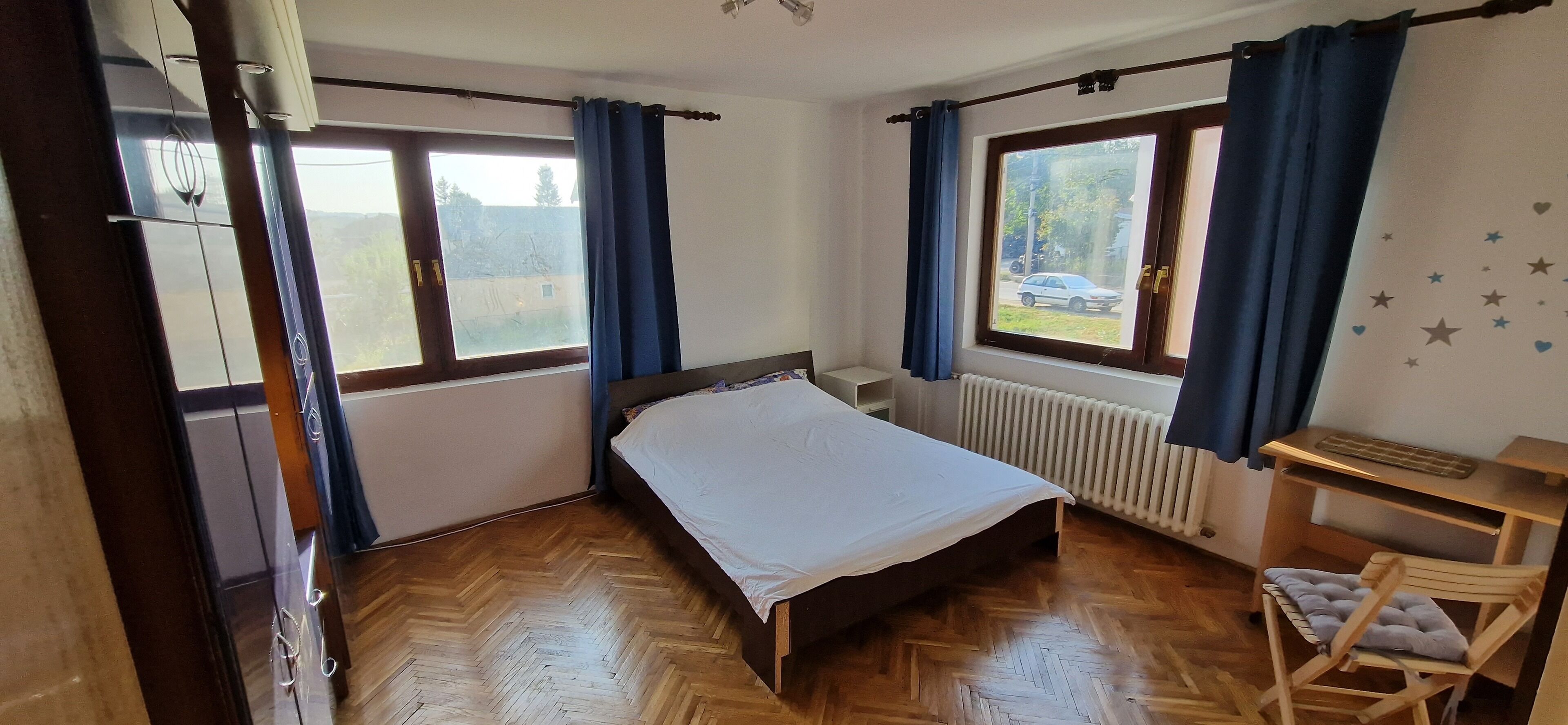 Une chambre avec un lit, un bureau et une chaise. Il y a deux fenêtres avec des rideaux, un radiateur et une vue sur l’extérieur.