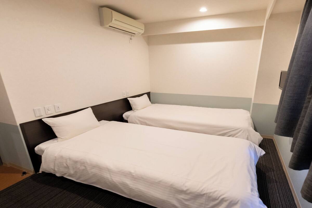 A hotel room with two single beds, a dark headboard, a wall-mounted air conditioner, and a curtain on the right side.