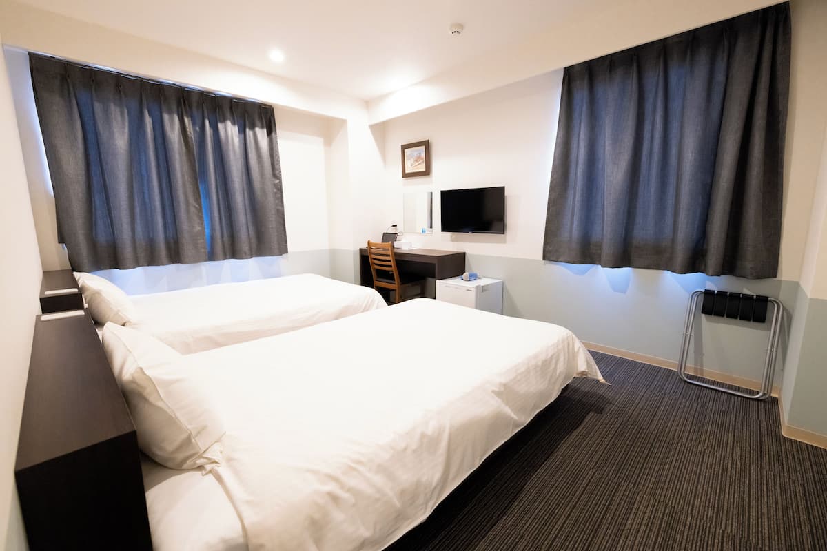 A hotel room with two beds, a desk, a chair, a TV, and a window with curtains.
