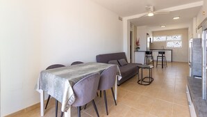 Ein modernes Wohnzimmer mit einer Couch, einem Esstisch und Stühlen. Es gibt eine Küchenzeile mit einer Arbeitsplatte und Hockern.