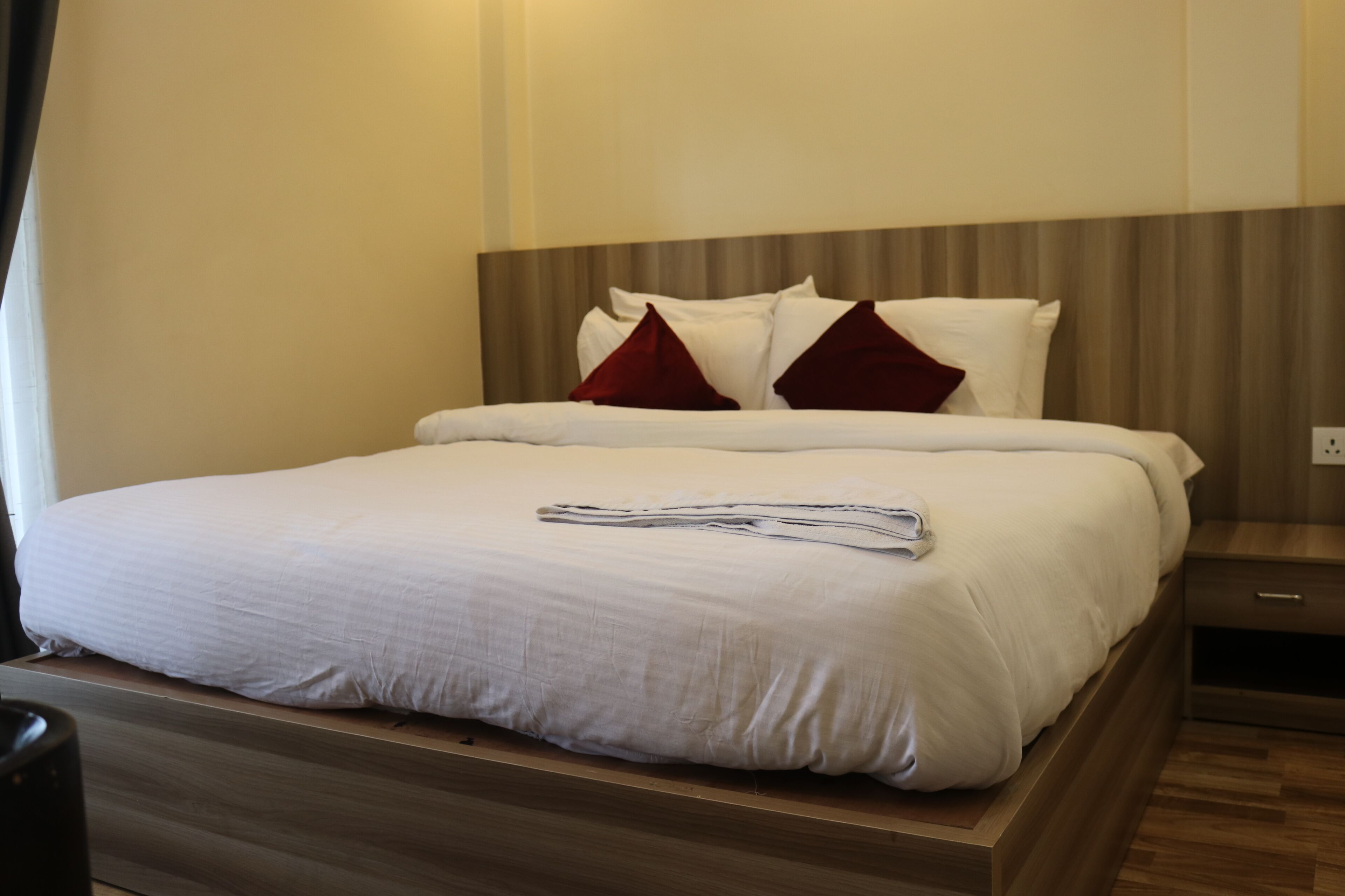 Una cama bien hecha con sábanas blancas y dos almohadas rojas, una mesita de noche de madera con cajón, y un enchufe en la pared.