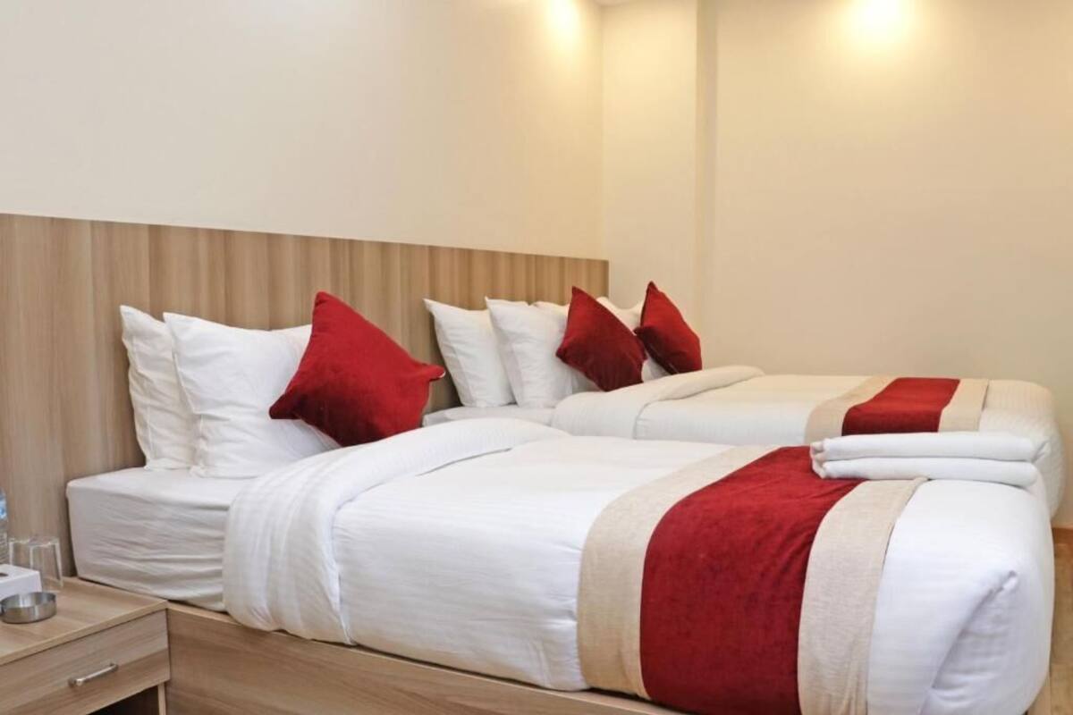 A hotel room with two beds, each with white bedding and red pillows. There is a wooden headboard, a bedside table with a lamp, and a small drawer.