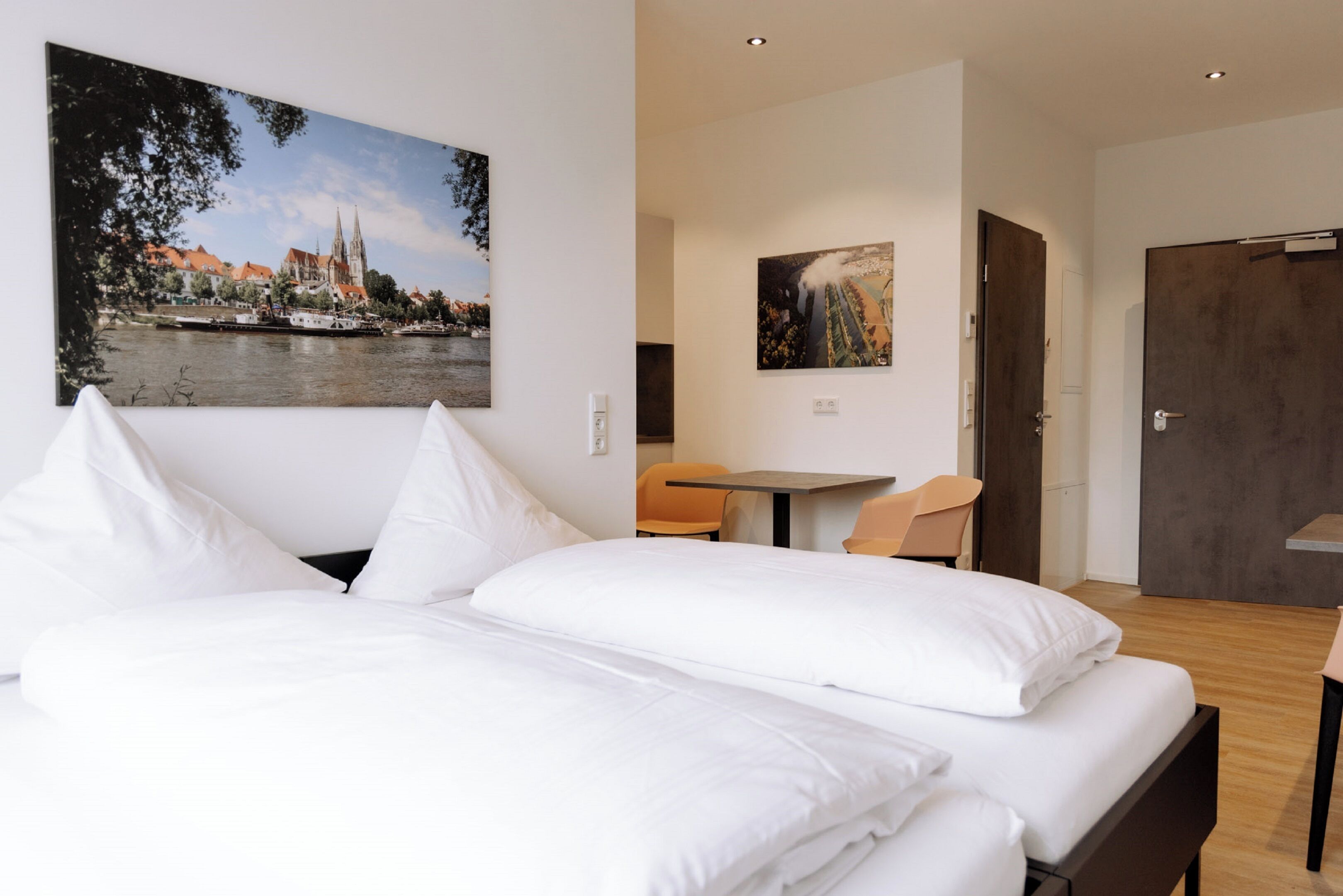 A hotel room with a bed, a desk, and two chairs. There are paintings on the walls and a door to the right.