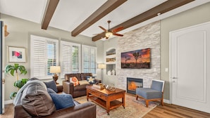 A living room with a fireplace, a ceiling fan, and a large window with shutters. - LV370 Vibrant Downstairs 3BR Legacy Villa Lock-Off (La Quinta)