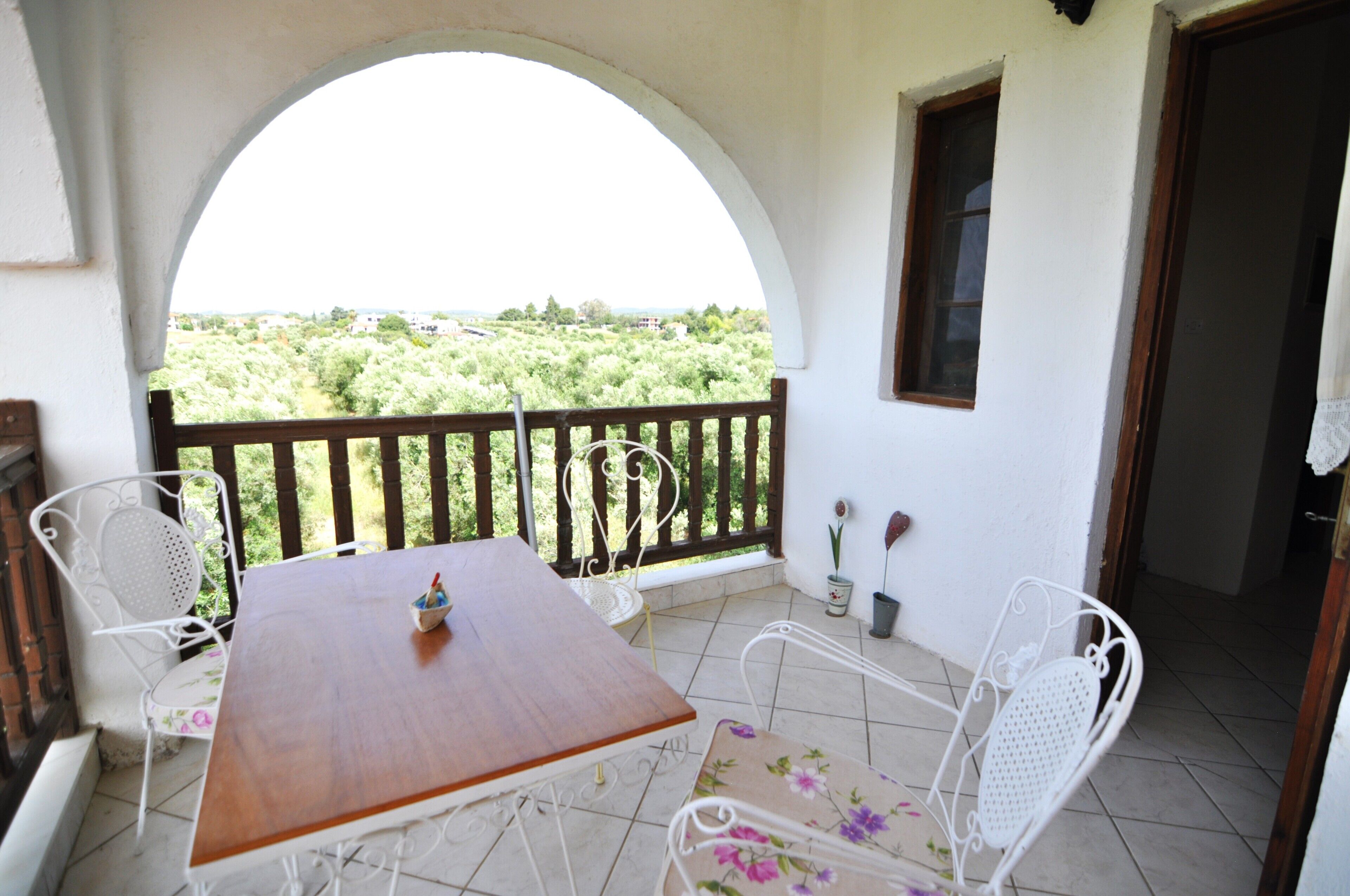 Un balcón con mesa de madera y sillas de metal blanco, con vista a un paisaje verde y exuberante.