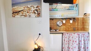 Ett kök med en platt-TV som är monterad ovanför en bänkskiva, ett litet bord med en lampa och en vägghylla med köksartiklar.