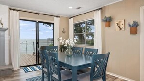 Uma sala de jantar com mesa e cadeiras, um vaso com flores e vista para o oceano através de uma porta deslizante.
