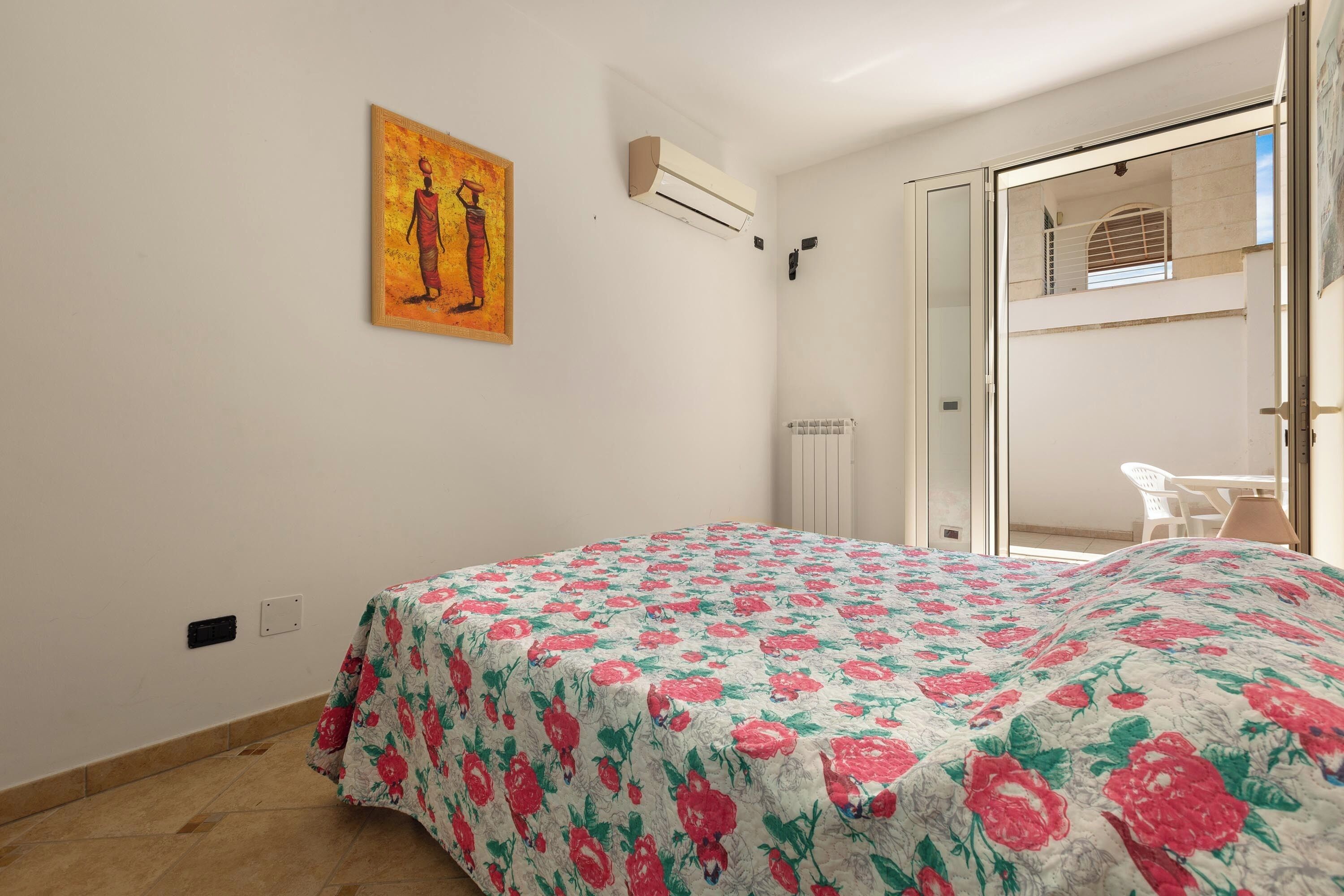 A bedroom with a floral bedspread, a painting on the wall, and a view of a balcony with a chair.
