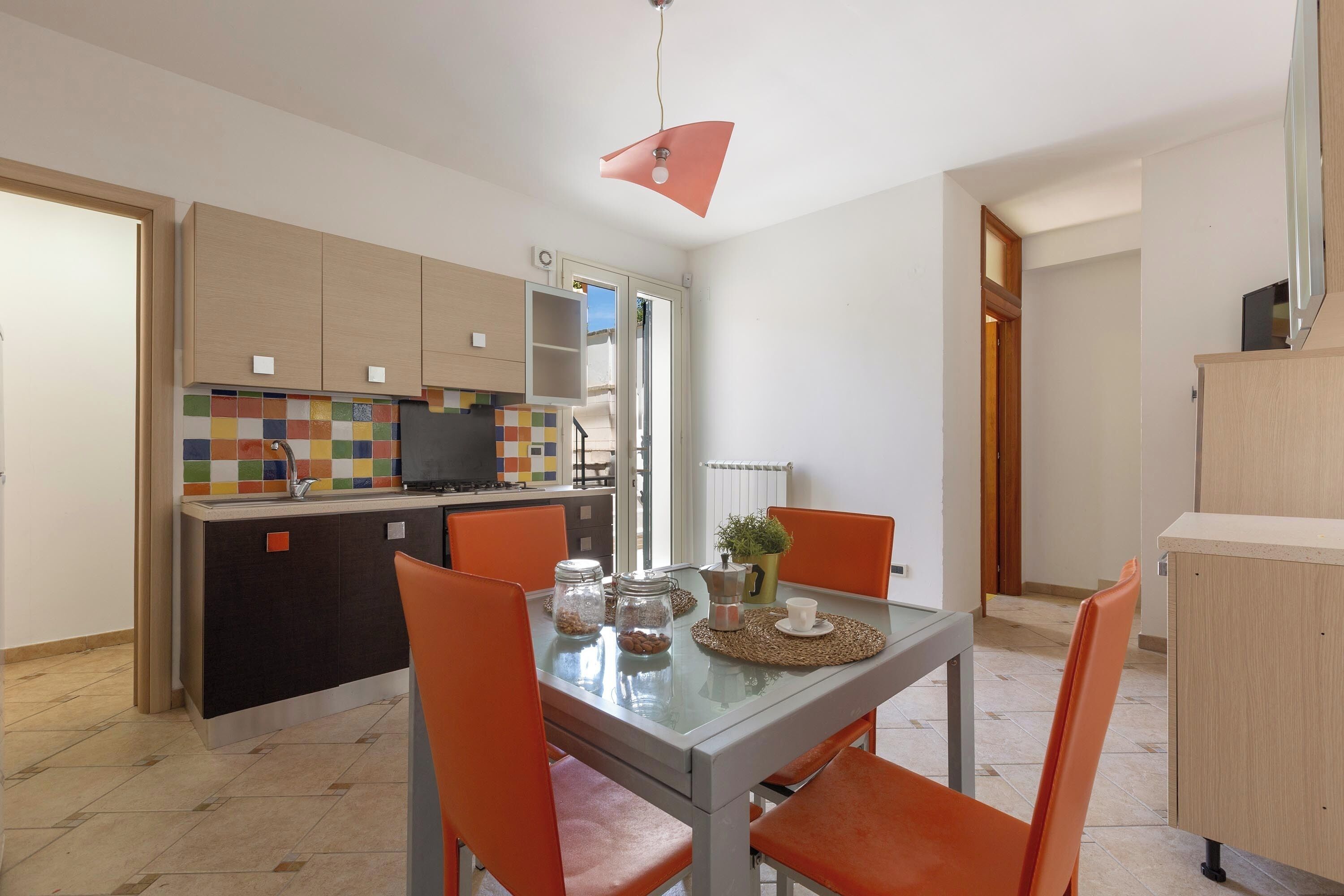 Una cocina moderna con un salpicadero colorido, un comedor con sillas naranjas y una pequeña mesa con una cafetera y frascos.