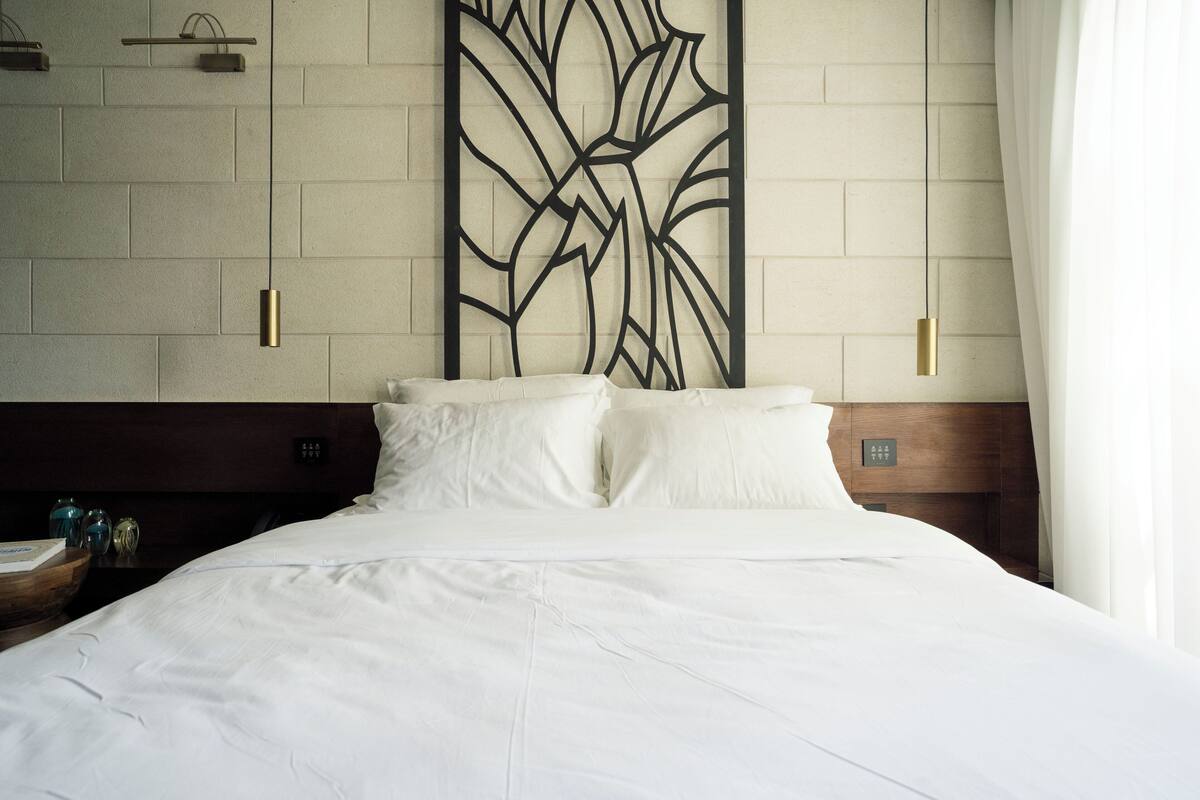 A hotel room with a large bed, a wooden headboard, a decorative wall panel, and a nightstand with a lamp.