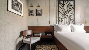 A hotel room with a large bed, a wooden nightstand, a chair, and a patterned rug. There are framed pictures on the wall and a decorative wall panel.