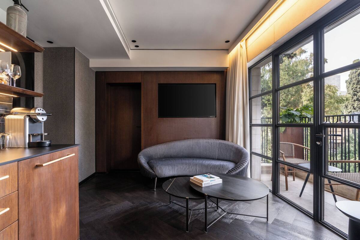 A modern living room with a dark wood floor, a round coffee table, a grey upholstered sofa, a flat-screen TV mounted on a wooden wall, and a large window offering a view of the outdoors.