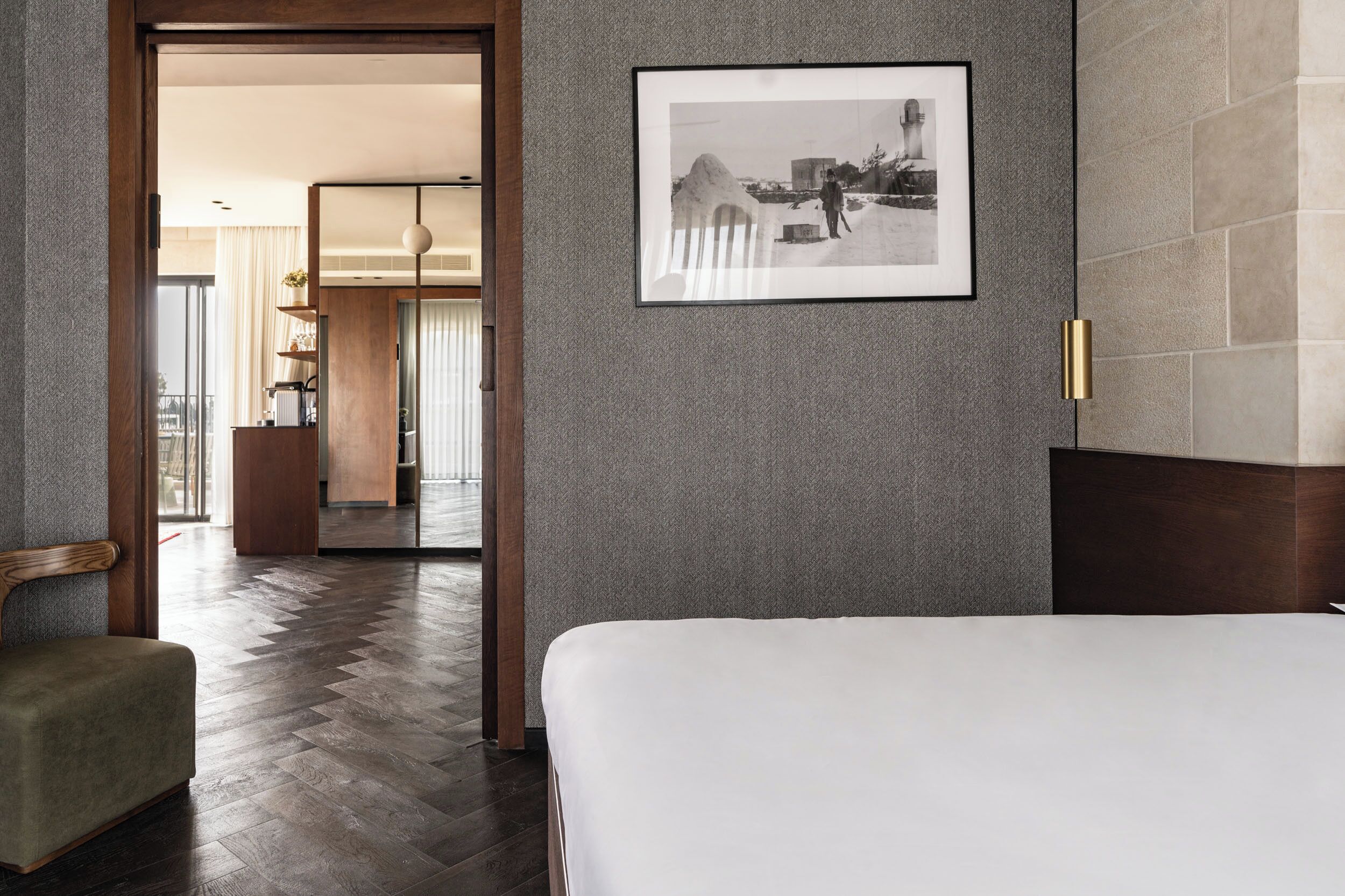 A hotel room with a large bed, a wooden chair, a framed picture on the wall, and a view of the hallway and balcony.