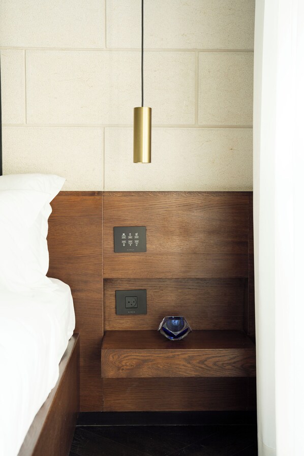A hotel room with a wooden headboard, a bed with white bedding, a bedside table with a blue object, and a wall-mounted light fixture. - Ramban 20 Hotel Jerusalem (Jerusalem)
