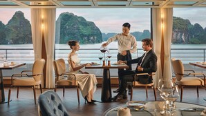 Une salle à manger avec une table dressée pour quatre personnes, un serveur qui sert du vin et une vue sur une côte montagneuse.