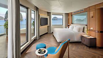 A modern hotel room with a large bed, a TV, a small table with a cake, and a view of the sea.