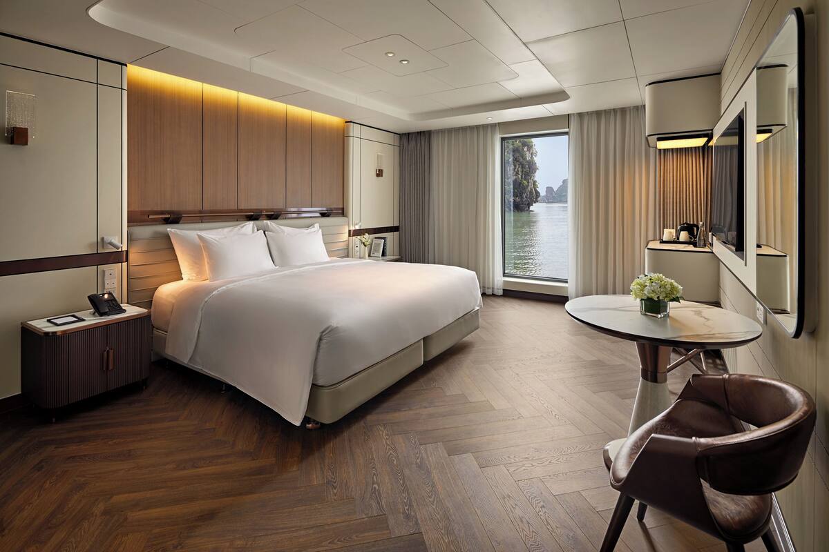 Ein modernes Hotelzimmer mit einem großen Bett, einem Nachttisch mit Lampe, einem runden Tisch mit Stuhl und Blick aufs Wasser.