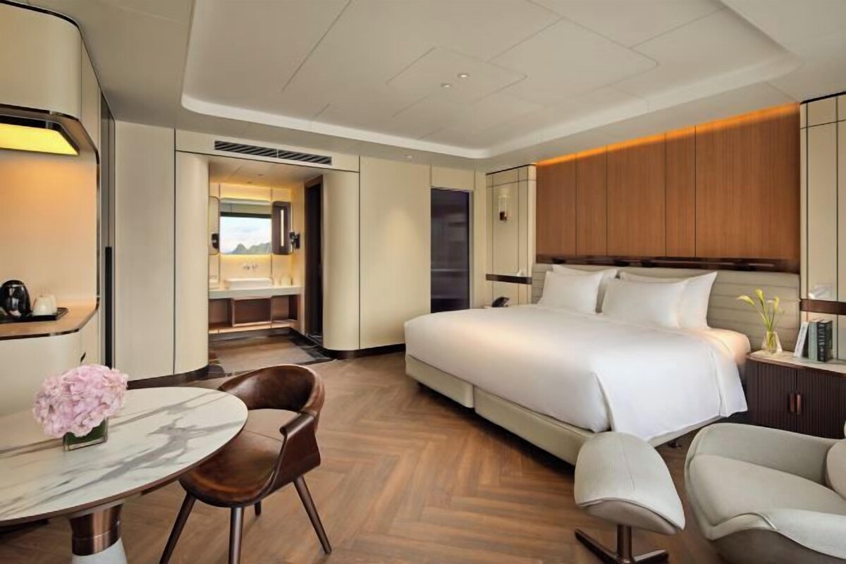 Een moderne hotelkamer met een groot bed, een zithoek met een tafel en stoelen, en een uitzicht op de omgeving door een geopende deur.