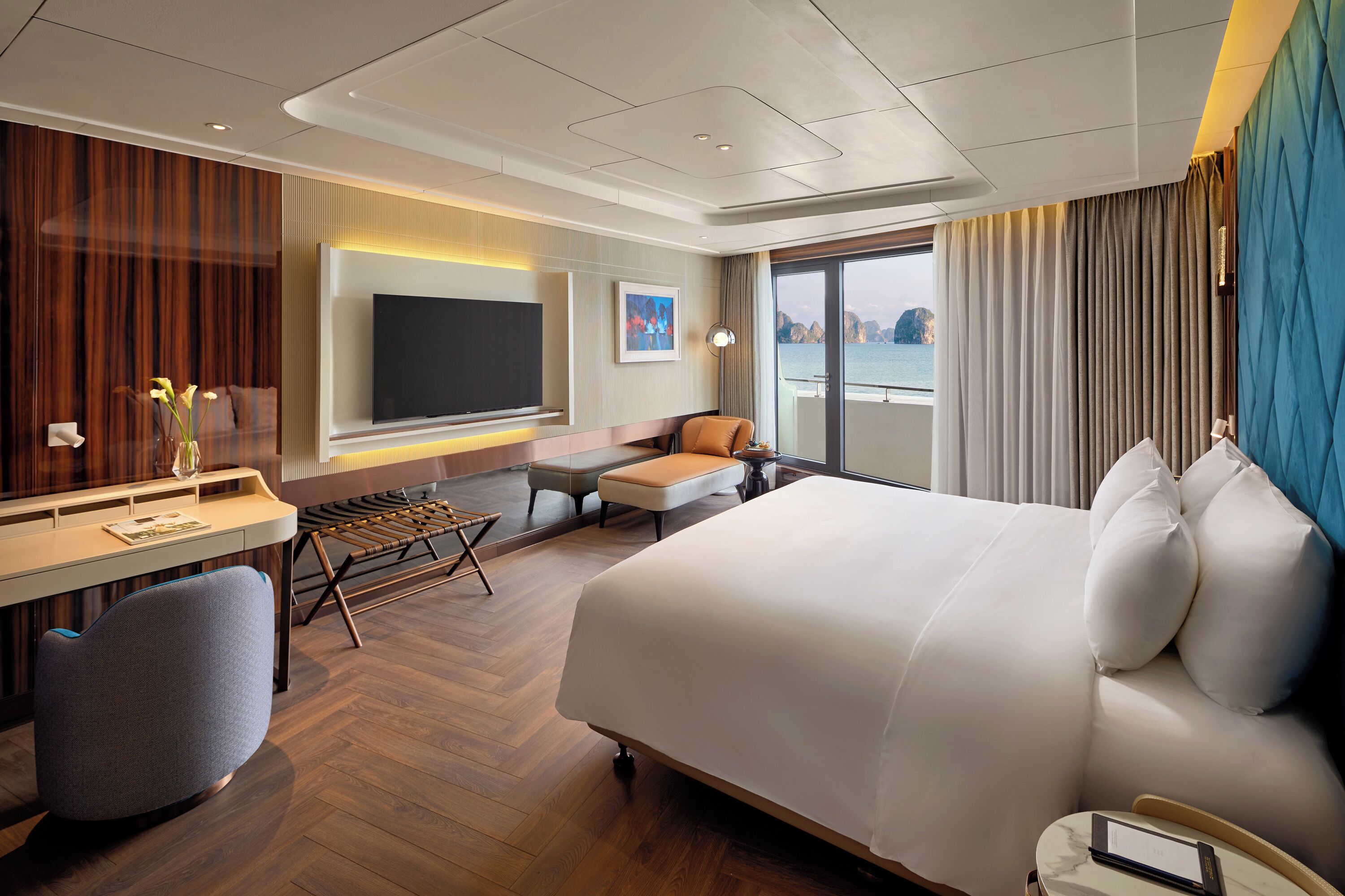 A hotel room with a large bed, a desk with a flat-screen TV, a seating area with a sofa and armchairs, and a view of the sea through a large window.