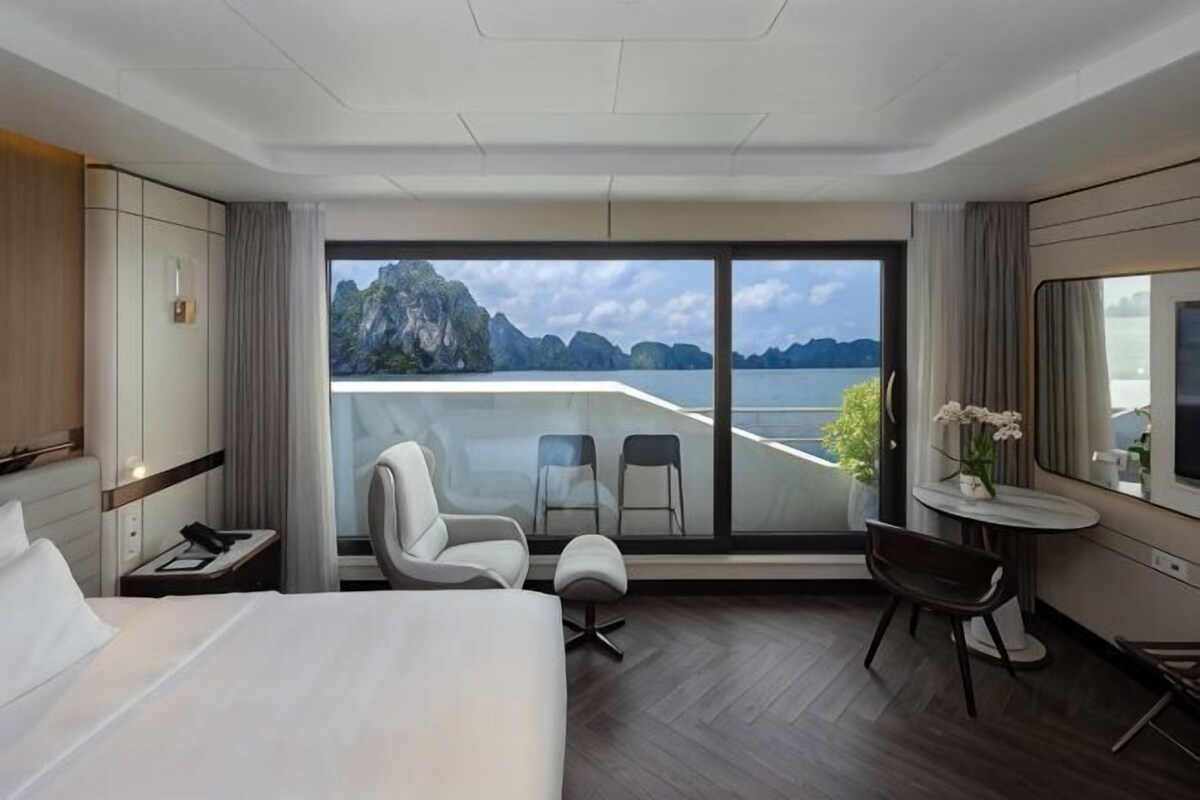 Ein Hotelzimmer mit Bett, Schreibtisch, Stuhl, einem kleinen Tisch mit Vase und einem großen Fenster mit Meerblick.