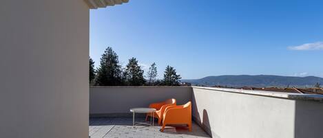 A rooftop terrace with a table and two chairs, a clear view of trees and distant hills.