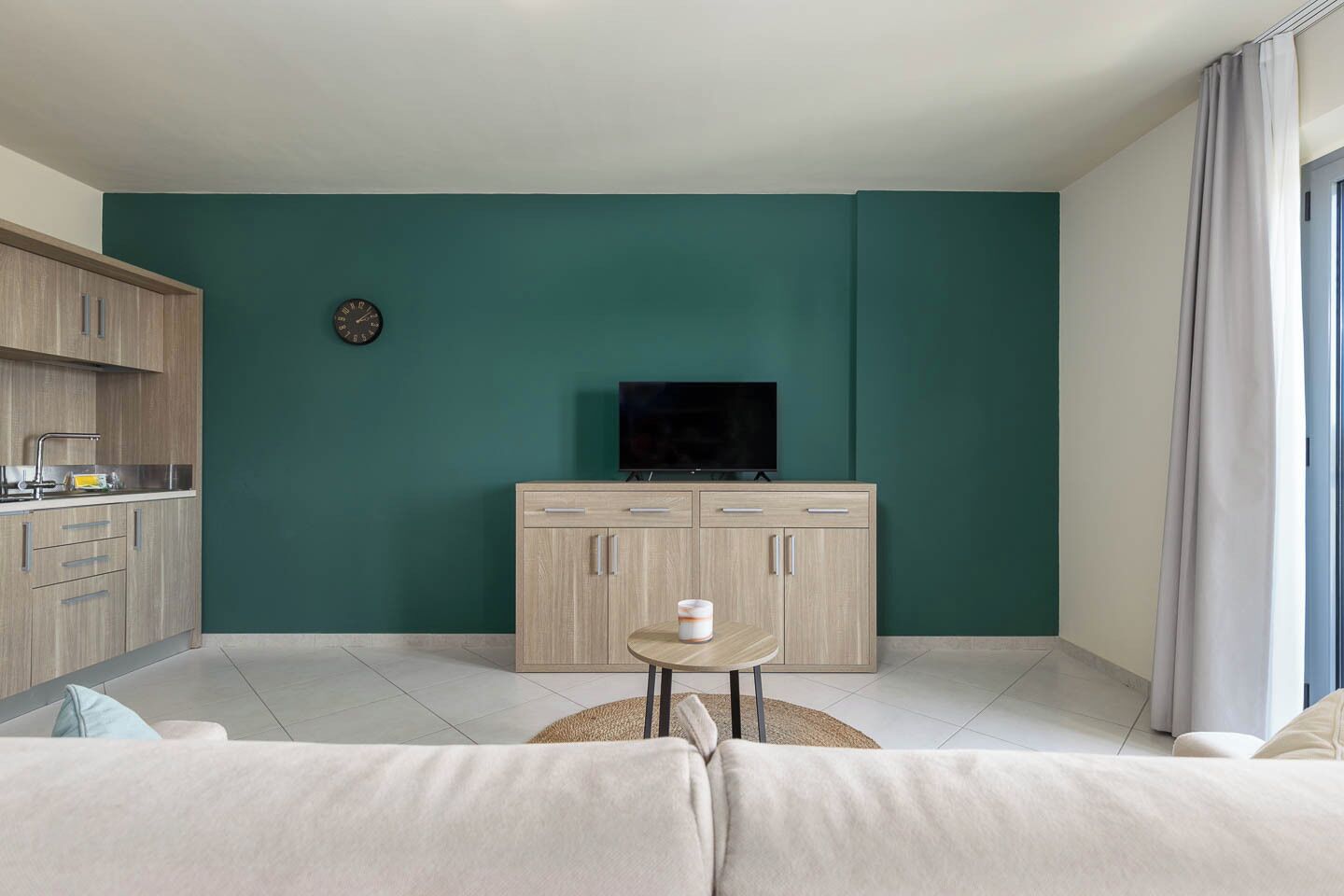 Een moderne woonkamer met een turquoise accentmuur, een flatscreen televisie, een houten kast, een klein rond tafeltje met een kaars en een bank.
