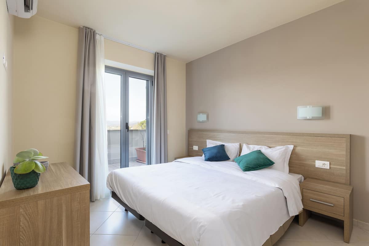 A hotel room with a bed, a bedside table with a plant, a nightstand, and a view of the outdoors through a sliding door.
