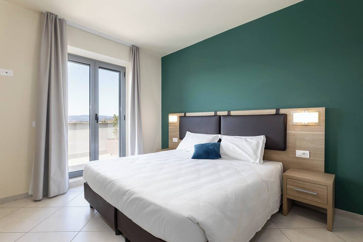 A hotel room with a large bed, a nightstand, a wall-mounted lamp, and a view through a sliding glass door.