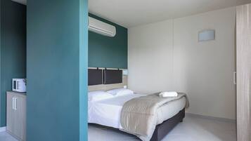 A modern hotel room with a bed, a built-in air conditioner, a small cabinet, and a door.