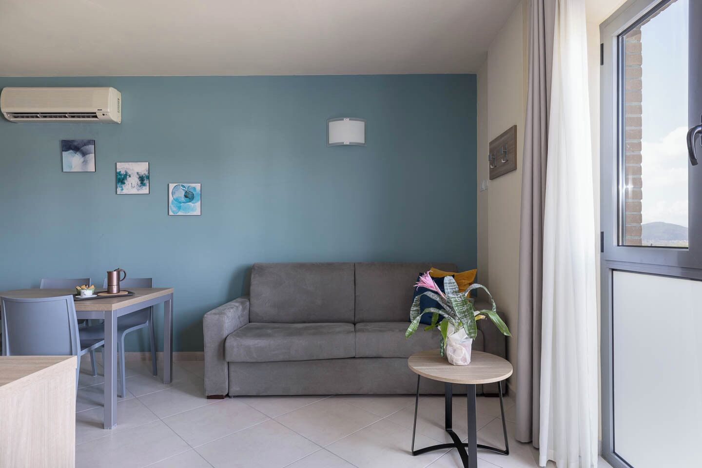 Ein Wohnzimmer mit grauem Sofa, einem kleinen runden Tisch mit Pflanze und einem Essbereich mit Tisch und Stühlen.