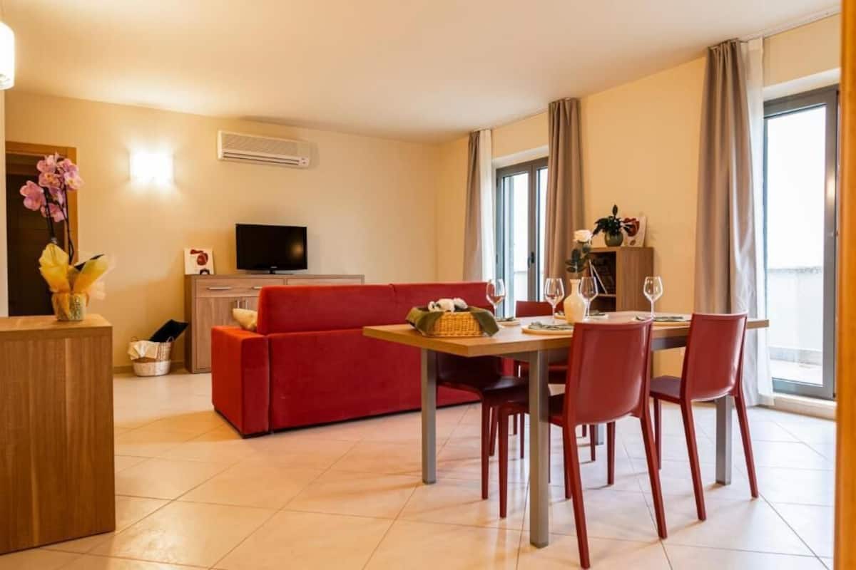 A modern living room with a red sofa, a dining table with four chairs, a flat-screen TV, and a large window with curtains.