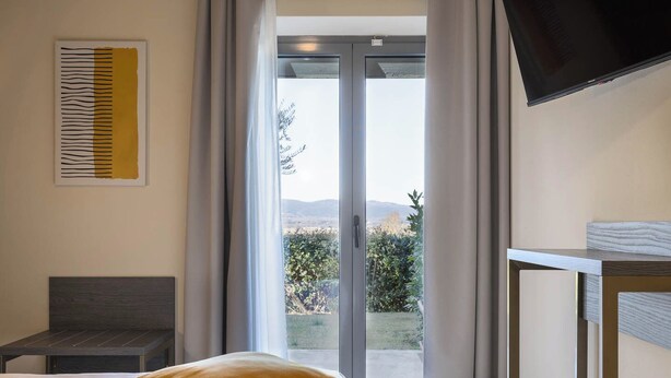 Una camera da letto con un letto, un comodino, una TV montata a parete e una porta scorrevole in vetro che dà su un balcone con vista su vegetazione e colline.