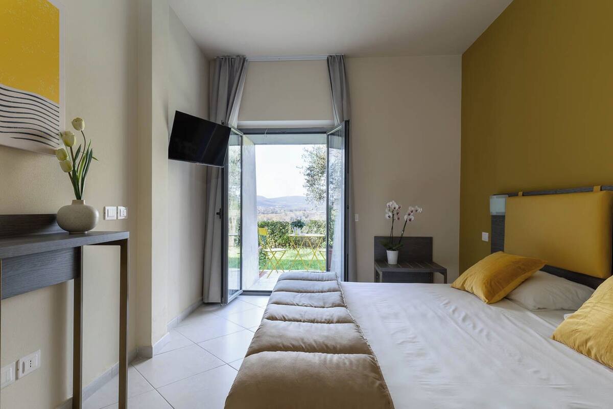 A bedroom with a bed, a bedside table, a TV mounted on the wall, and a view of the outdoors.