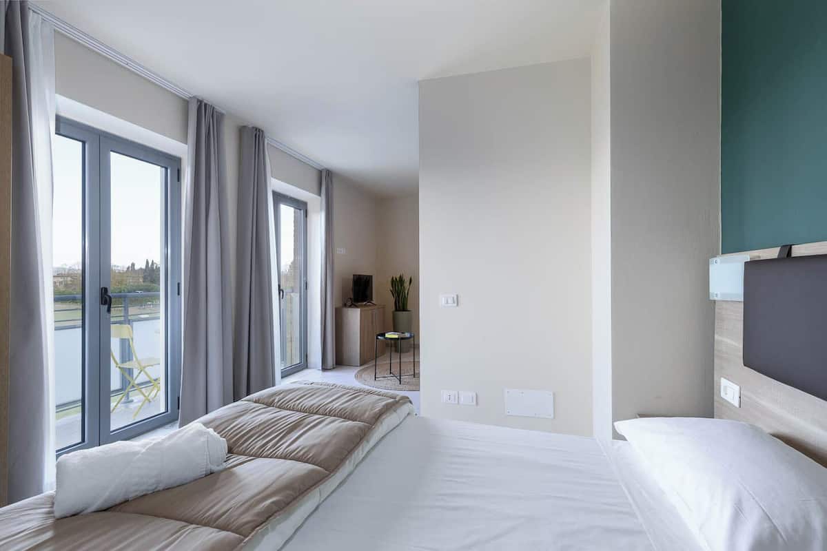 A hotel room with a large bed, a TV, a desk, and a view of the outdoors through the windows.