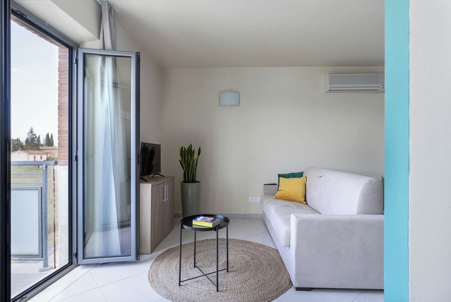 A modern living room with a sliding glass door leading to a balcony, a sofa with cushions, a small table with a book, a potted plant, and a wall-mounted air conditioner.