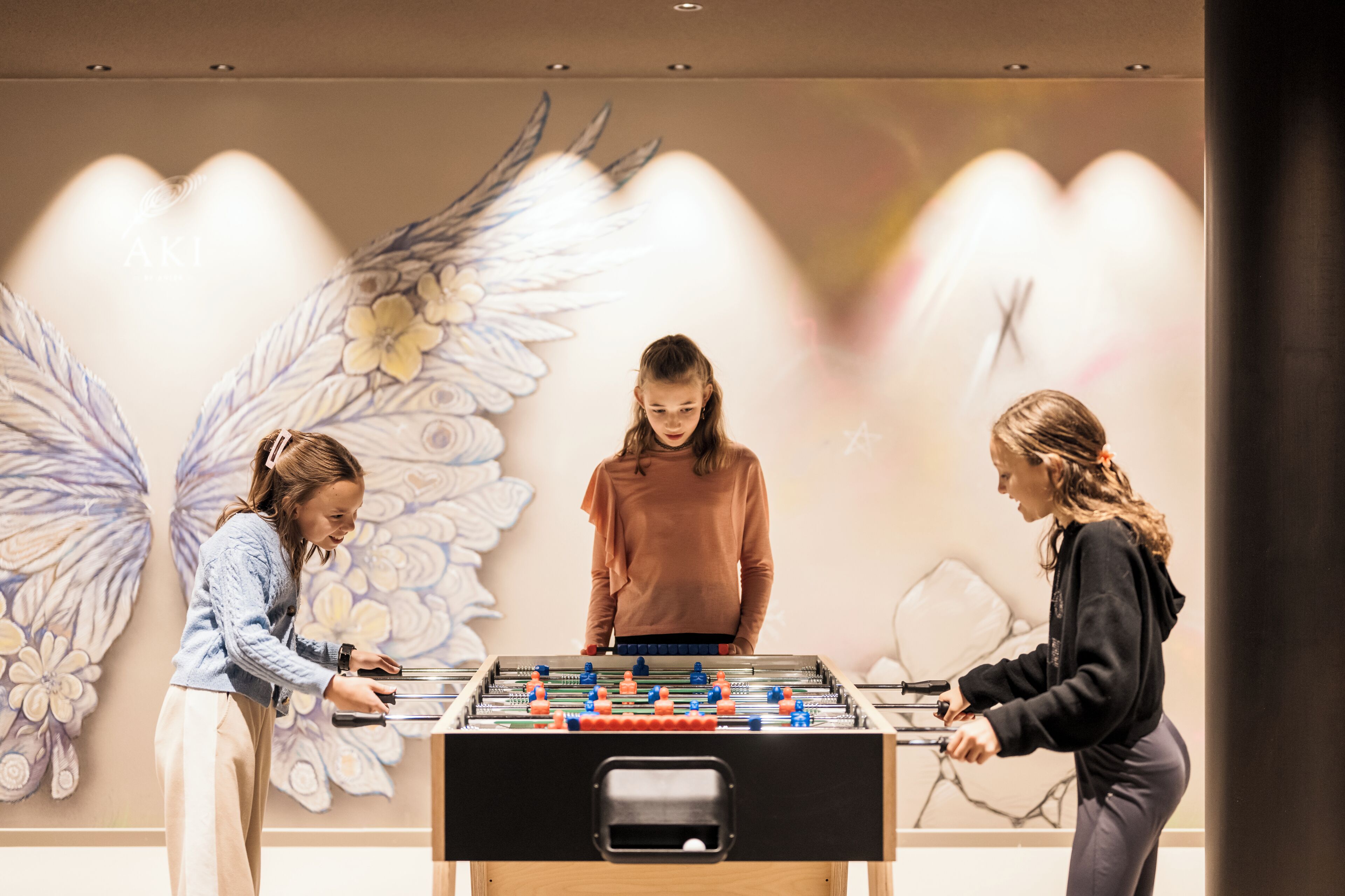 Three people playing foosball in a room with a large wall mural.