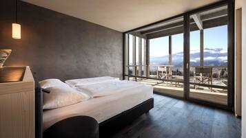 Une chambre moderne avec un grand lit, une vue sur les montagnes par une porte-fenêtre coulissante et une terrasse en bois à l’extérieur.
