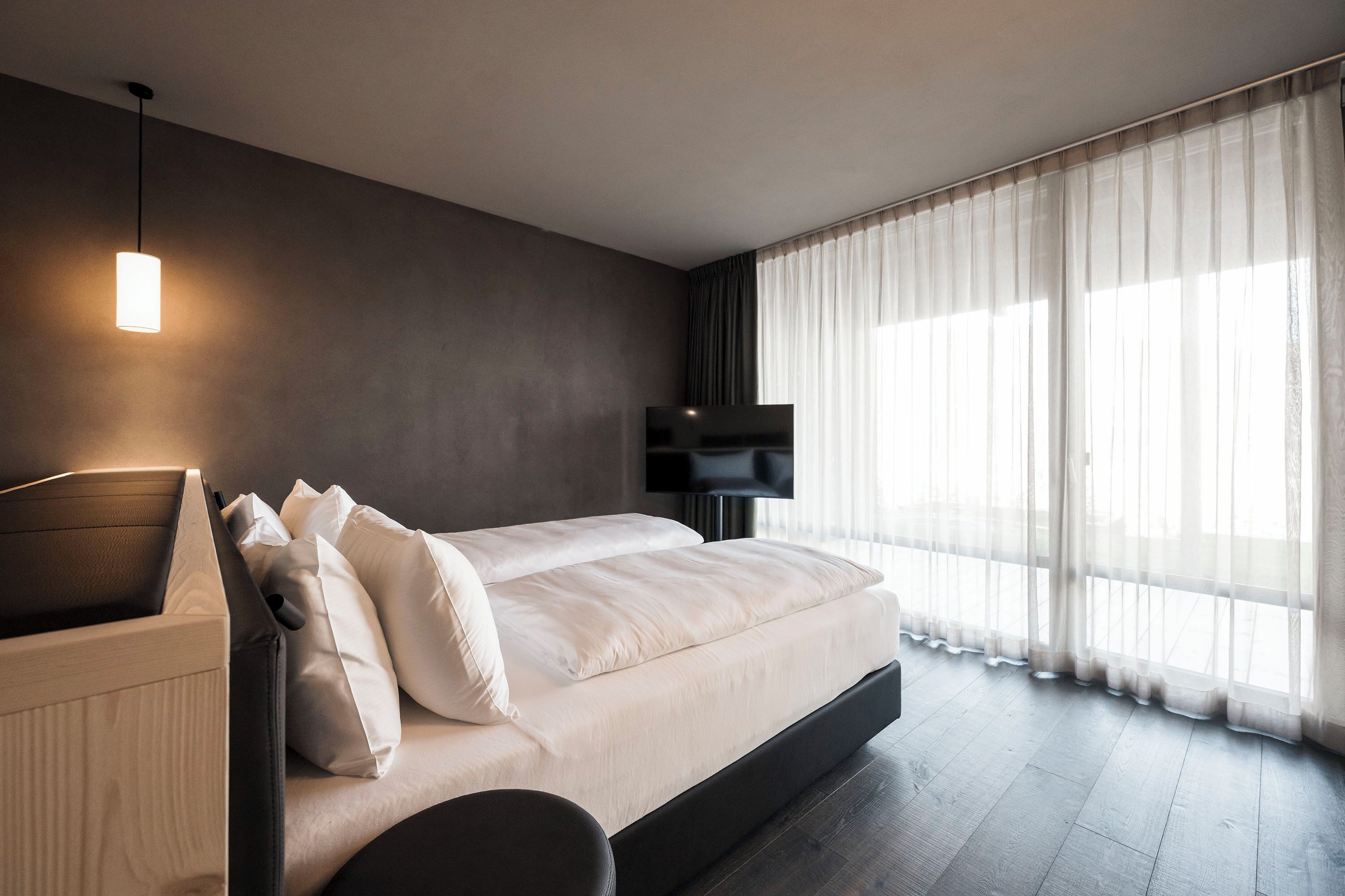 Ein modernes Schlafzimmer mit einem großen Bett, einem Nachttisch, einem Fernseher und transparenten Vorhängen.