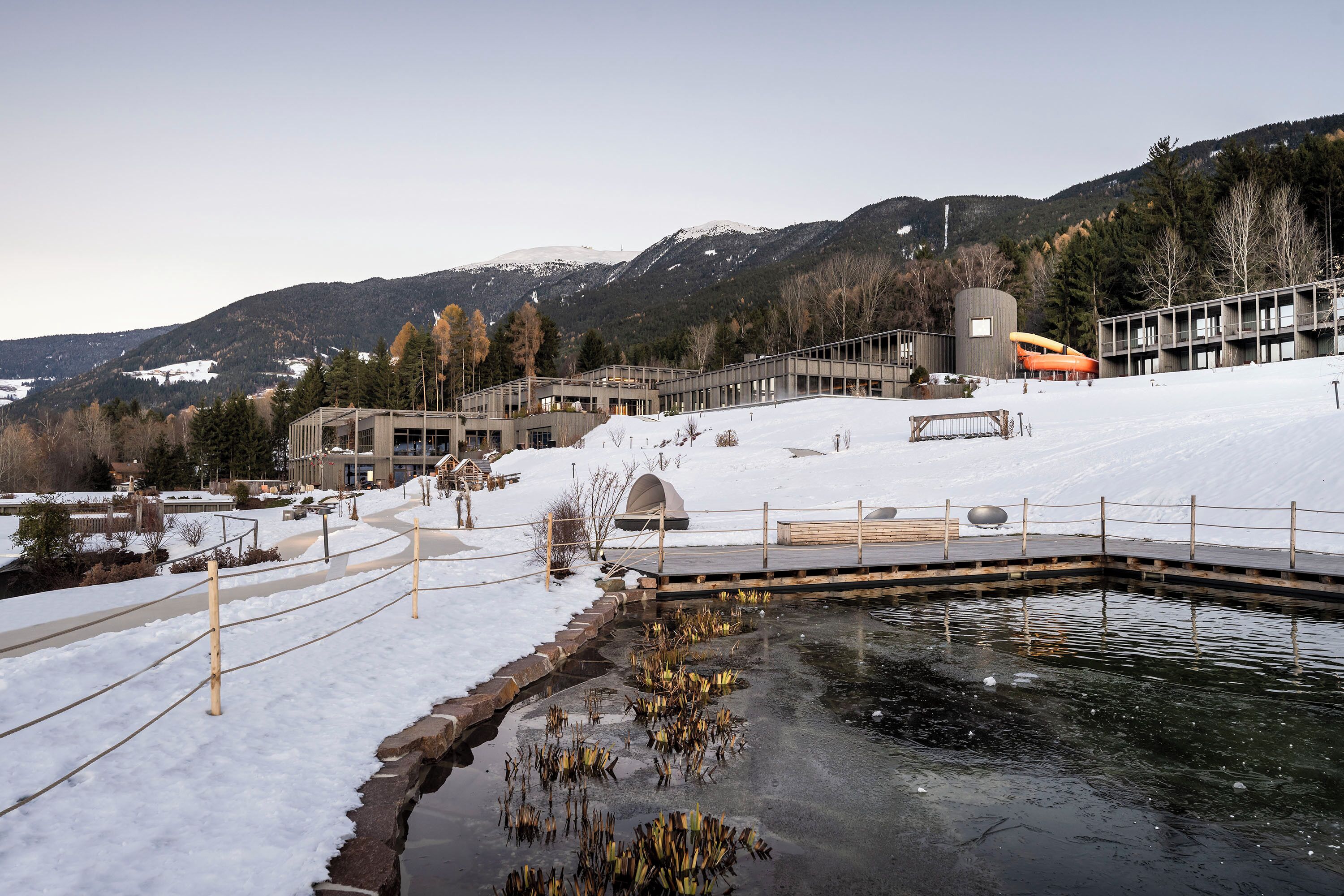A snow-covered outdoor area with a pond, a wooden deck, and a building with a modern architectural style.