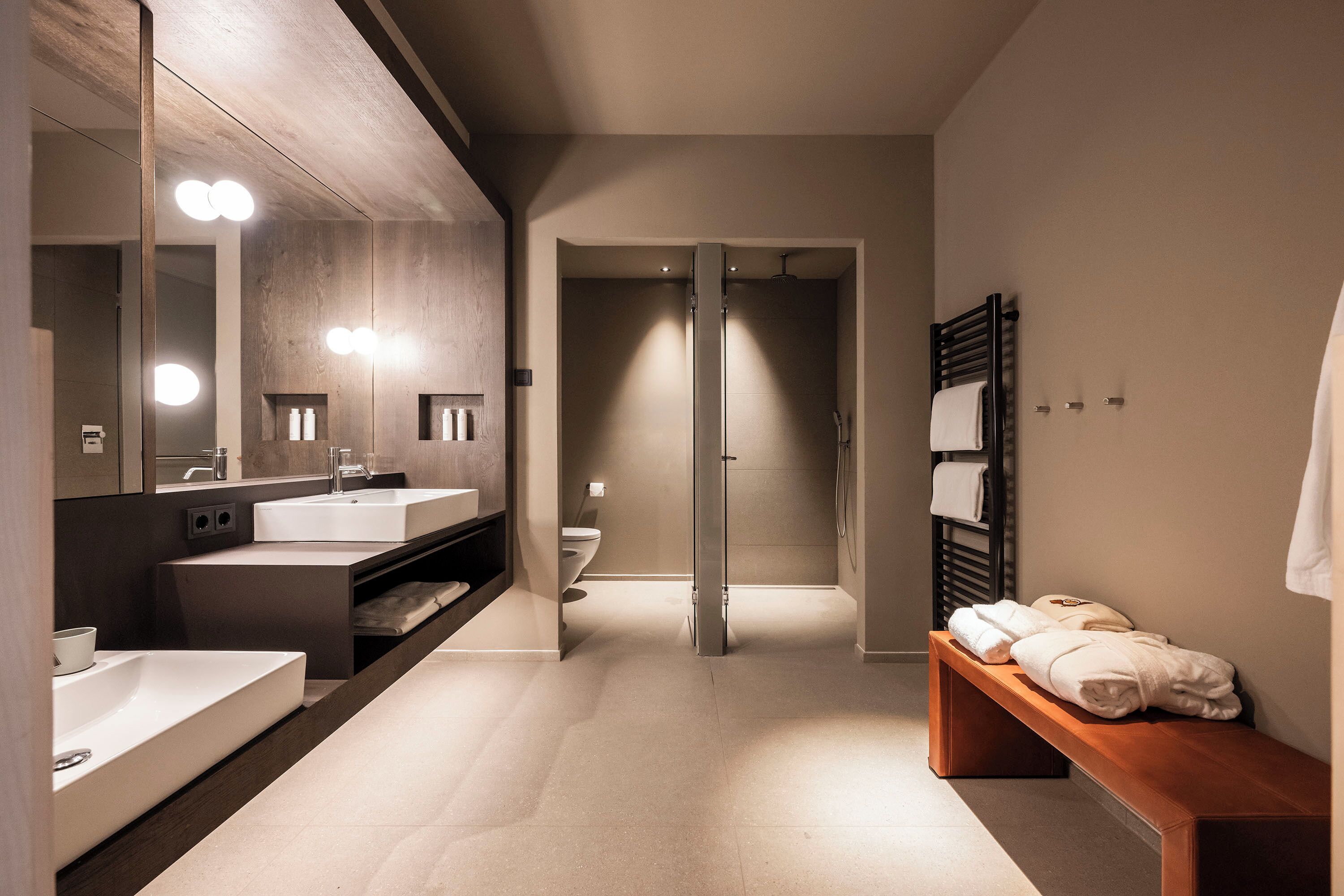 A modern bathroom with a large mirror, two sinks, a shower, and a bench with towels.
