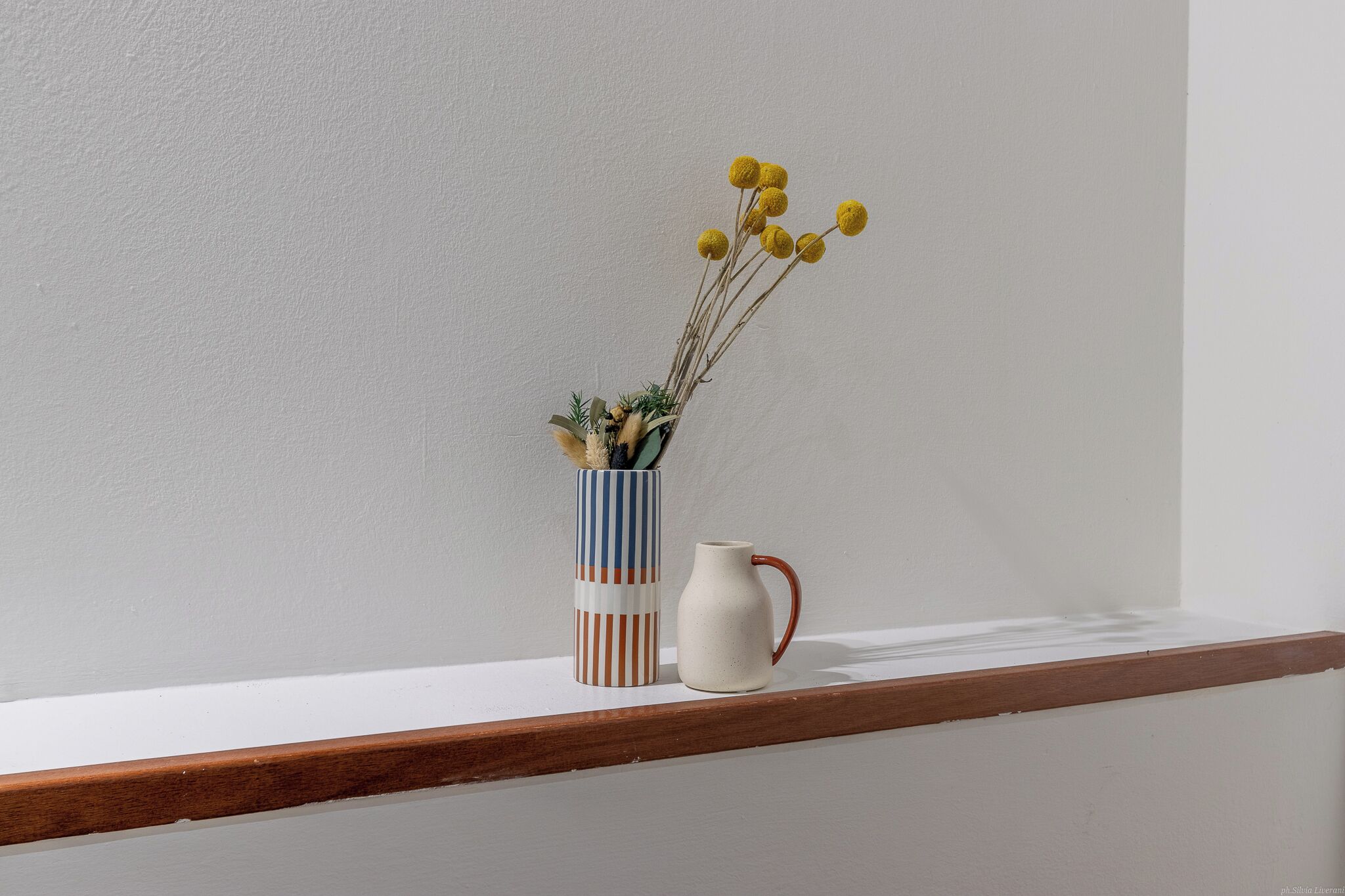 A white wall with a wooden shelf. On the shelf, there is a striped vase with dried plants and yellow flowers, and a white pitcher with a red handle.
