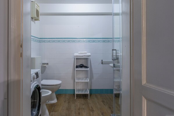 A compact bathroom with a washing machine, toilet, and a shelf with towels and toiletries. - HouSmart Moline 20 (Bologna)