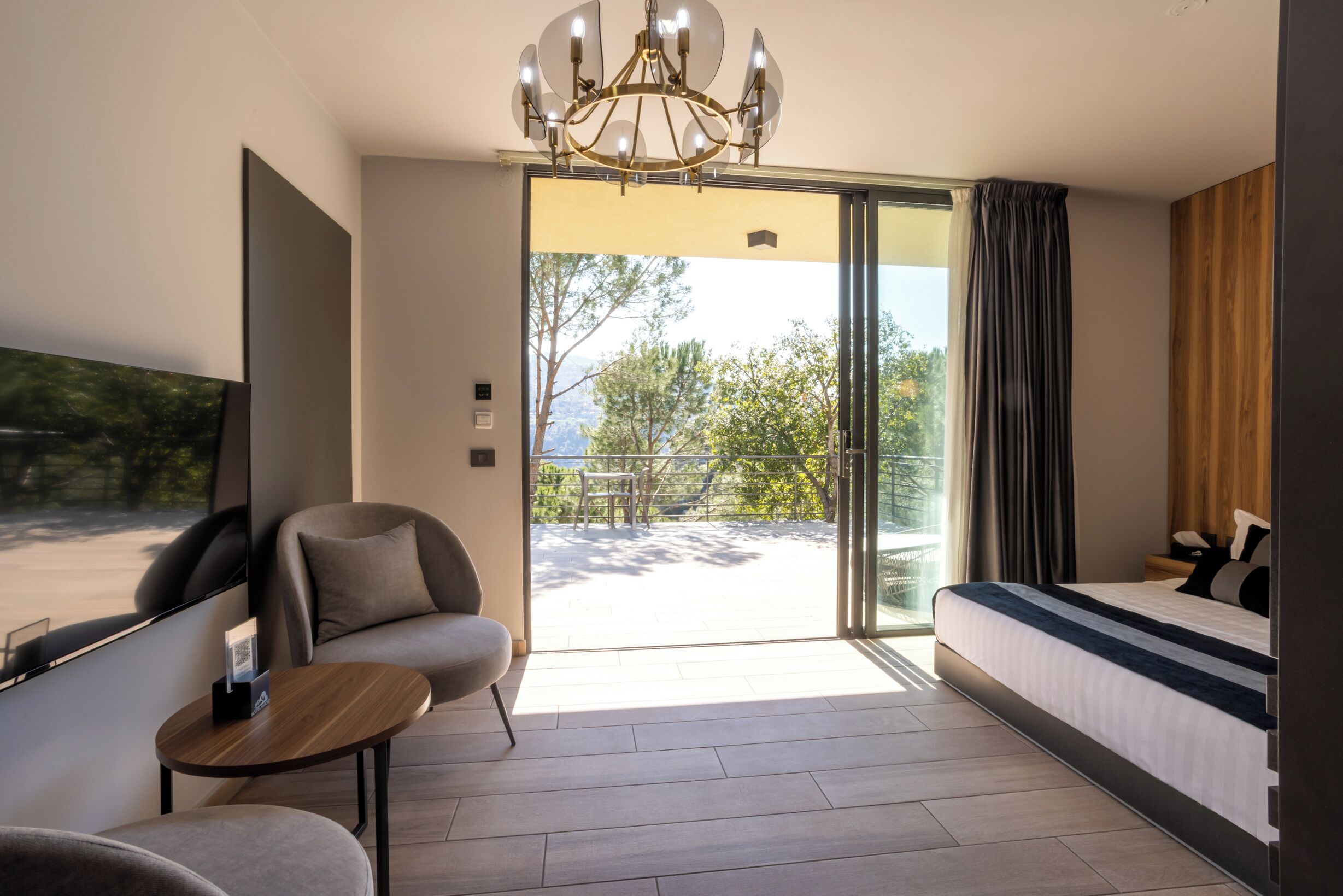 Habitación de hotel moderna con una cama grande, un sofá, una mesita y un televisor. La habitación tiene una puerta corrediza de vidrio que da a un espacio exterior con árboles y vistas al cielo.