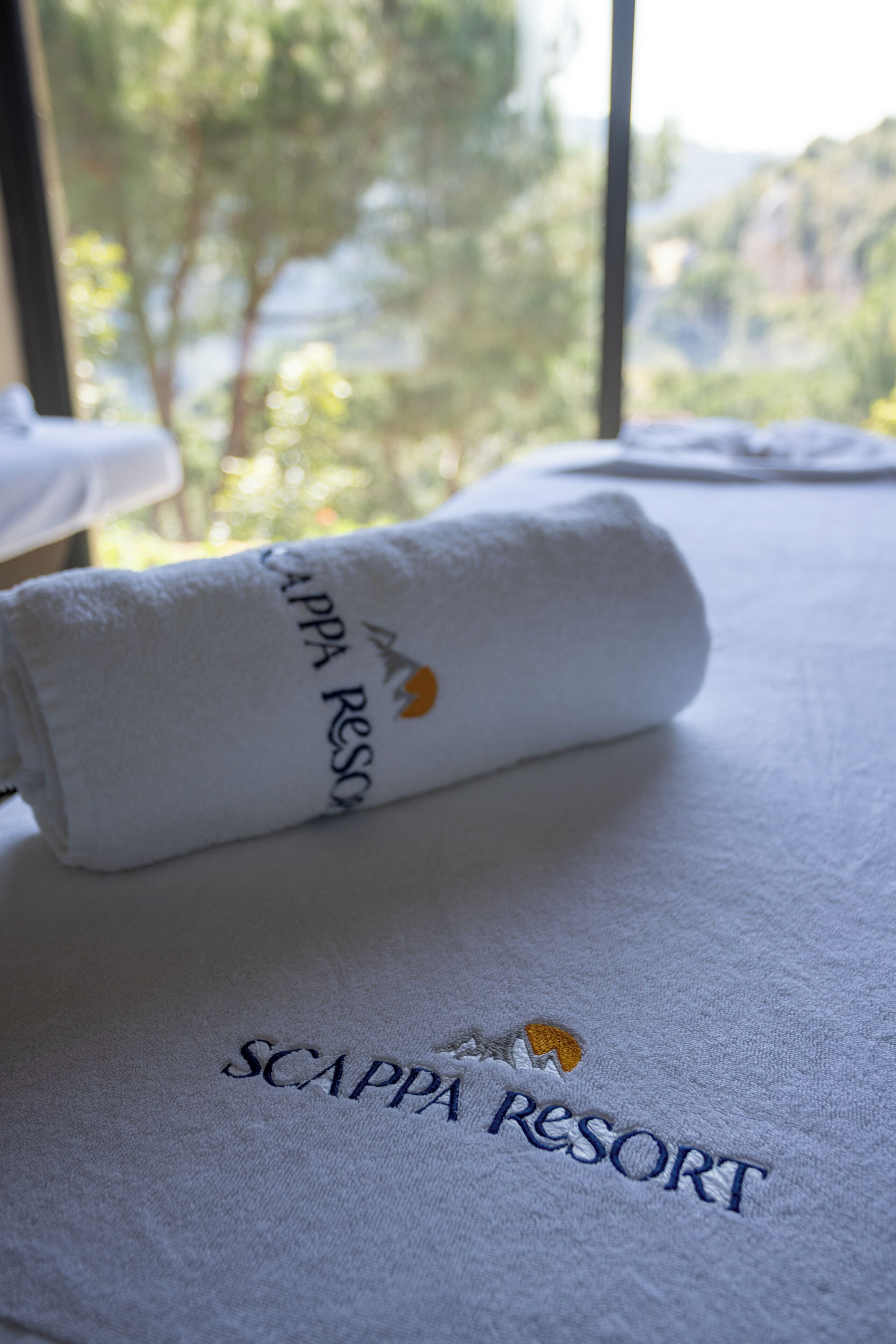 Una toalla blanca enrollada con el bordado "SCAPPA RESORT", sobre una cama con vista a árboles y montañas.