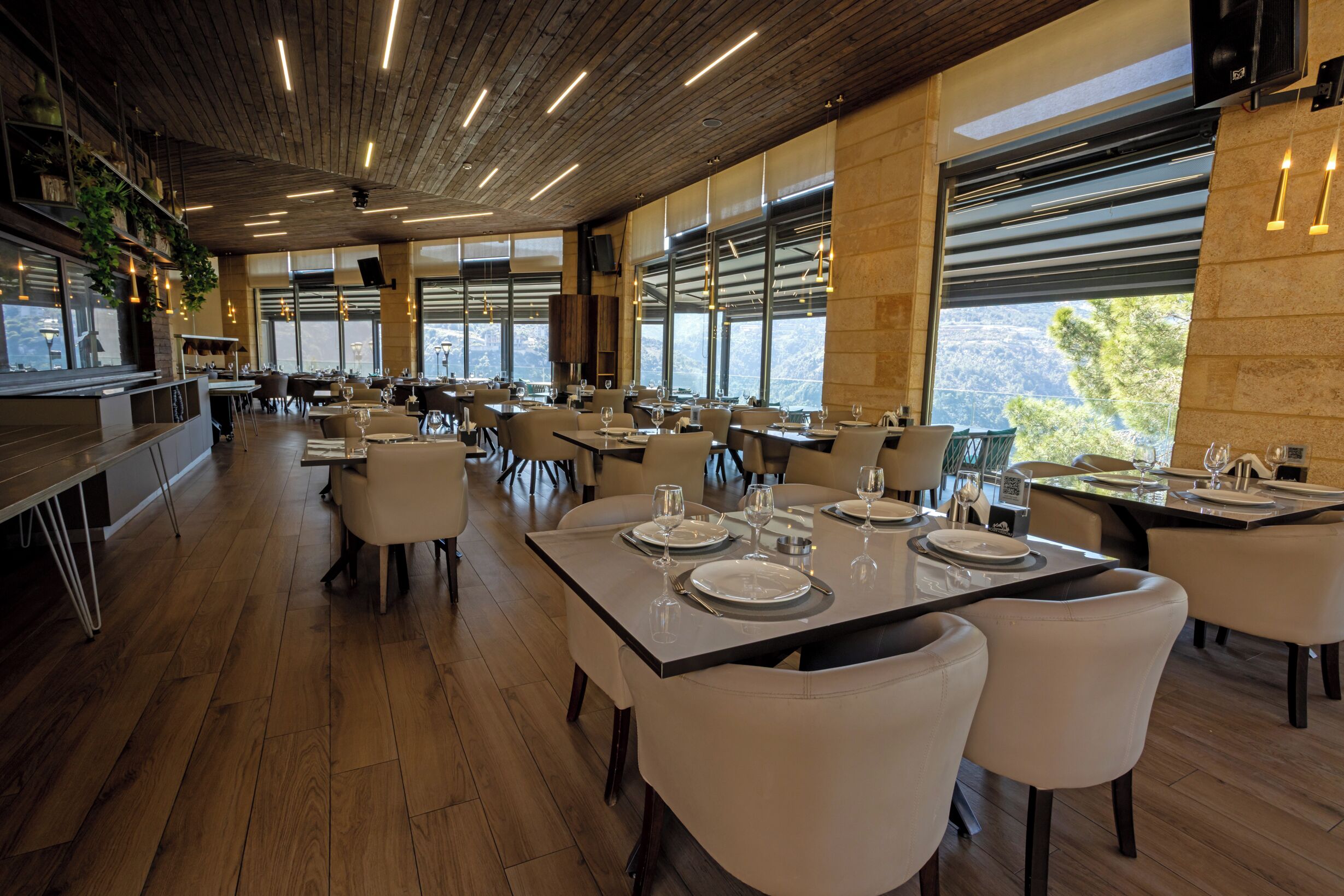 Un restaurant spacieux avec de hauts plafonds, un plancher en bois et de grandes fenêtres offrant une vue sur l’extérieur. Les tables sont dressées avec des nappes blanches et de beaux couverts.