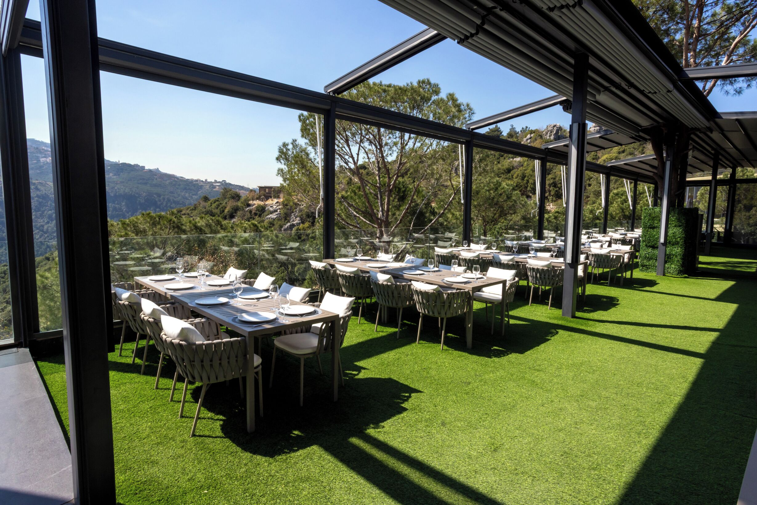 An outdoor dining area with tables and chairs, surrounded by glass walls and a view of a hilly landscape.