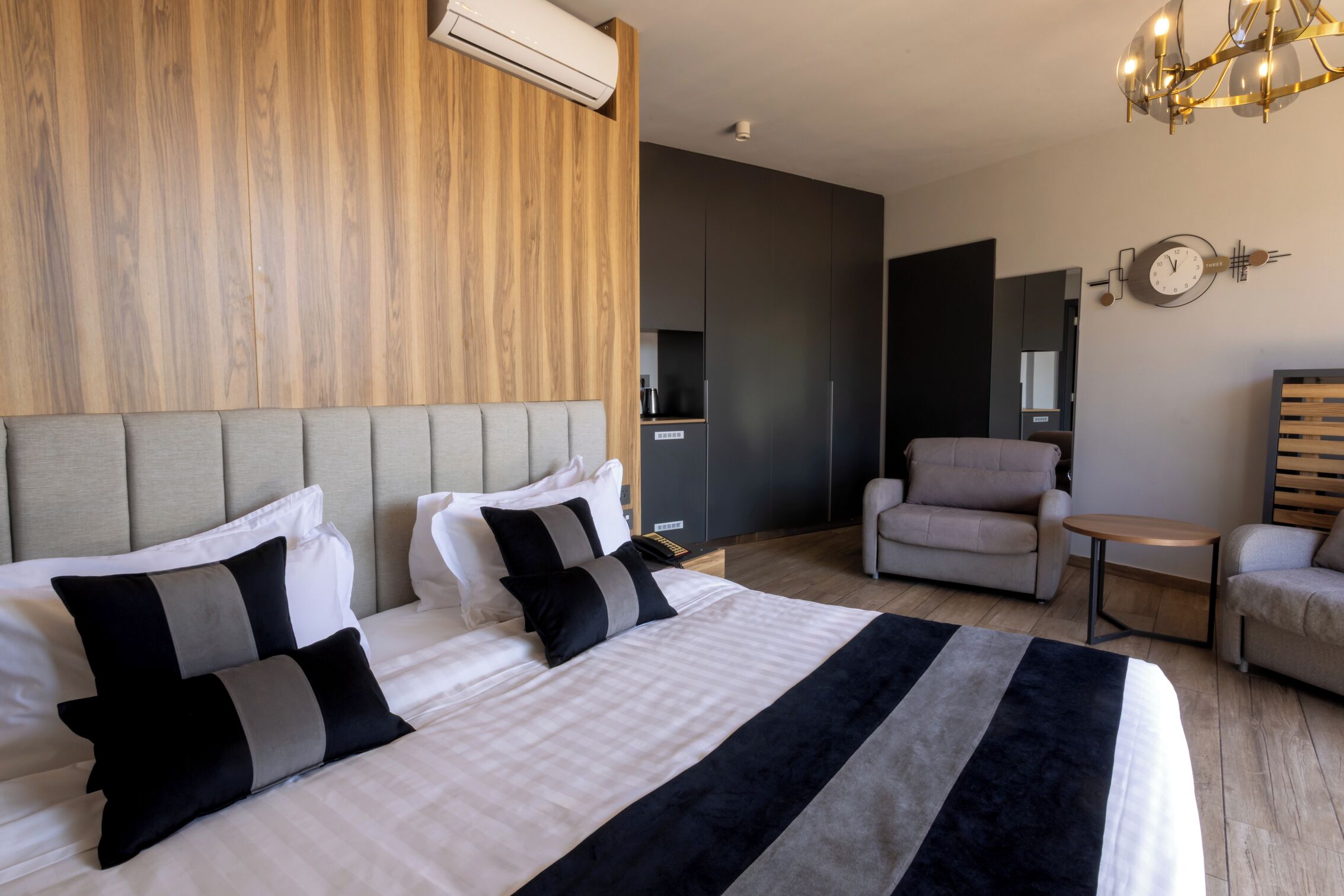 A modern hotel room with a large bed, a grey upholstered armchair, a small round table, and a wooden accent wall.