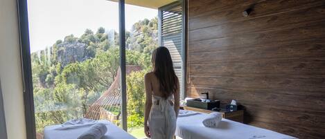 A spa room with two massage tables, a wooden ceiling, and a view of greenery outside.