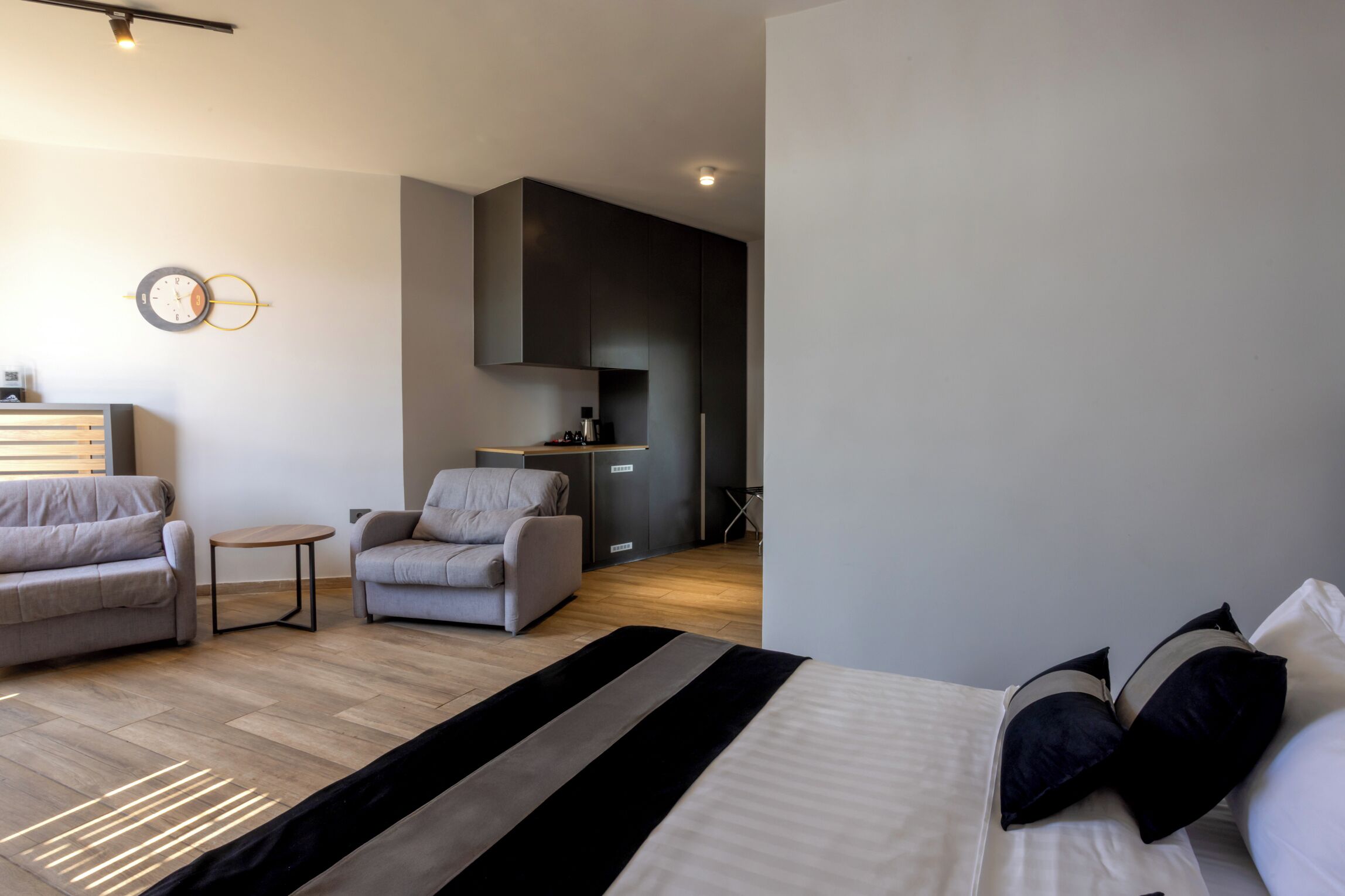 A modern hotel room with a bed, two sofas, a small table, and a kitchenette in the background.