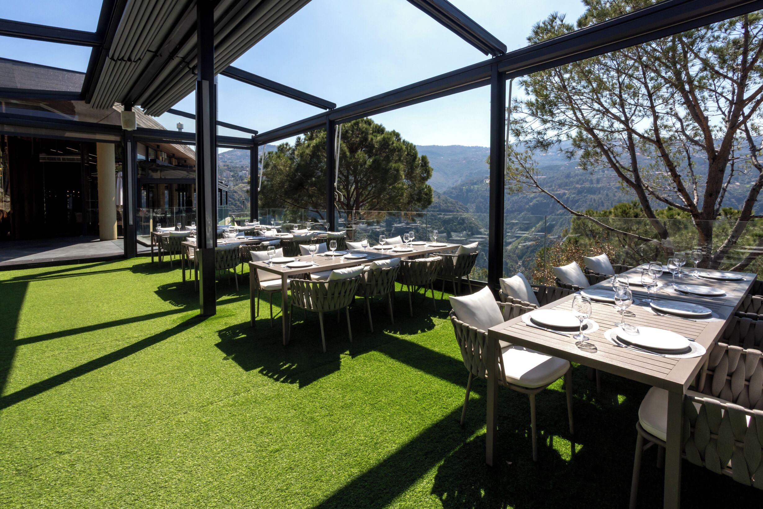 An outdoor dining area with tables set for two, surrounded by chairs, and a view of the landscape through large windows.